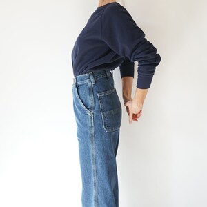 May include: A person wearing a navy blue sweatshirt and blue denim jeans. The jeans have a classic five-pocket design with a carpenter-style pocket on the right side. The outfit is simple and casual, suitable for everyday wear.