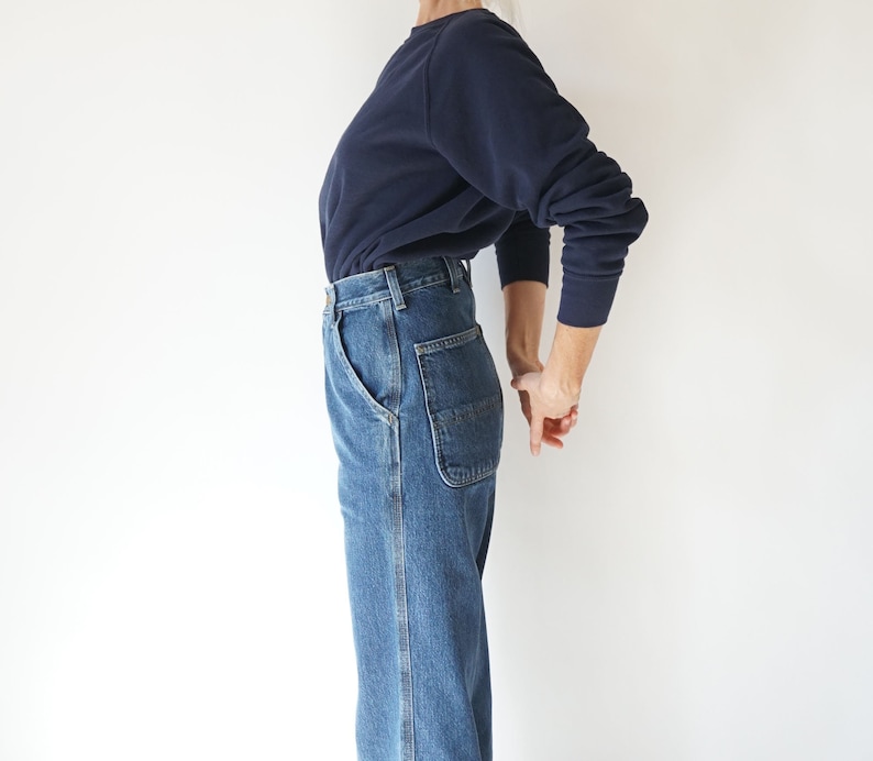 May include: A person wearing a navy blue sweatshirt and blue denim jeans. The jeans have a classic five-pocket design with a carpenter-style pocket on the right side. The outfit is simple and casual, suitable for everyday wear.