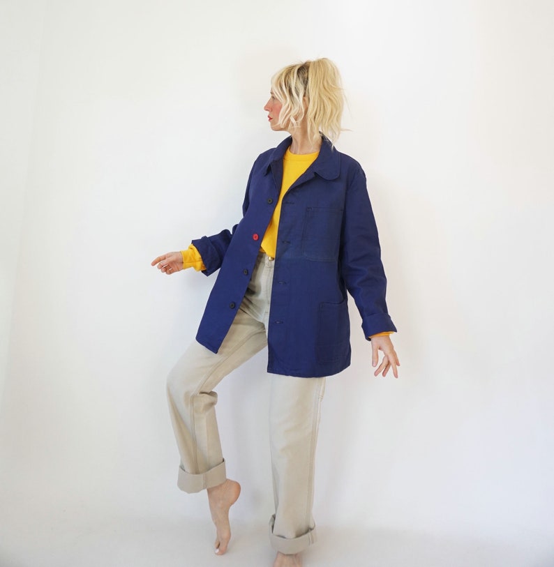 Pu&ograve; includere: Giacca blu navy con bottoni, due tasche frontali e un dettaglio con bottone rosso. Indossata sopra una camicia gialla a maniche lunghe e abbinata a pantaloni beige chiaro. L'outfit &egrave; ambientato su sfondo bianco.