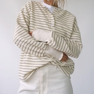 May include: Cream-colored striped long-sleeve shirt with a Henley neckline. The shirt features thin, dark stripes and is layered over a white long-sleeve shirt. The model is also wearing white pants.