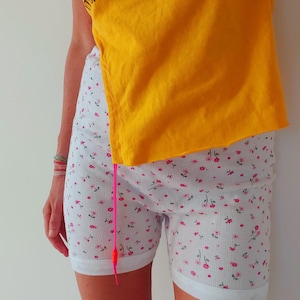 May include: White shorts with a pink floral pattern and a rolled hem. The shorts are paired with a yellow t-shirt. A pink drawstring hangs from the shorts.