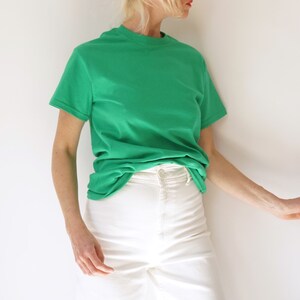May include: A bright green t-shirt with a crew neck and short sleeves. The shirt is paired with white denim pants. The model is leaning against a white wall. The outfit is simple and casual.