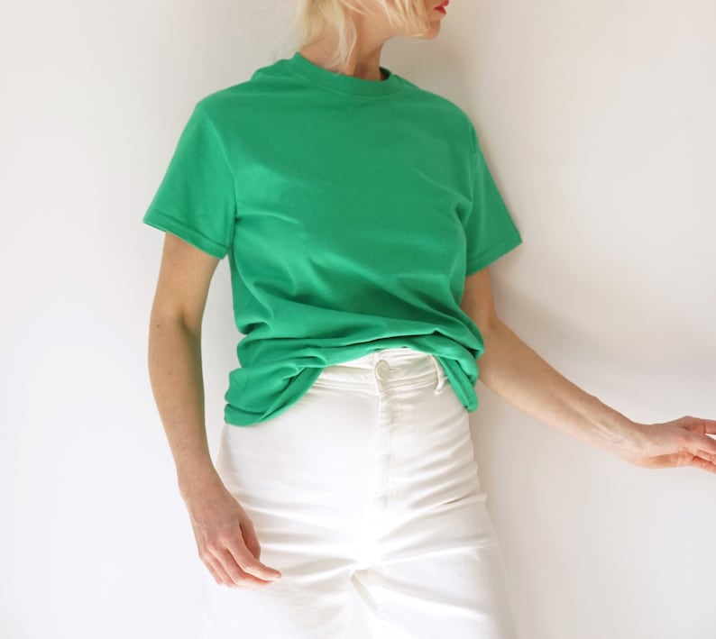 May include: A bright green t-shirt with a crew neck and short sleeves. The shirt is paired with white denim pants. The model is leaning against a white wall. The outfit is simple and casual.