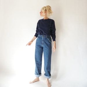 May include: A navy blue sweatshirt and blue denim jeans. The jeans have a high waist and are cuffed at the ankles. The model is wearing a casual outfit, perfect for everyday wear.