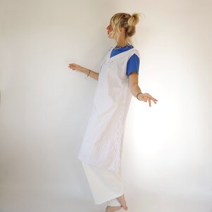 May include: A white and blue striped sleeveless dress with a V-neckline. The dress is worn over a blue t-shirt and white trousers.
