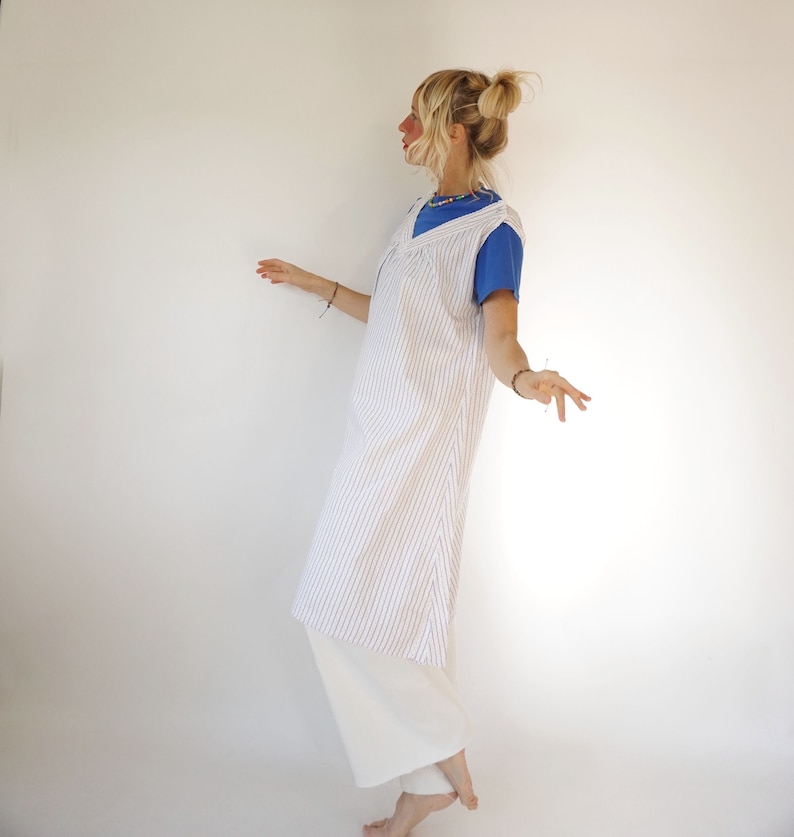 May include: A white and blue striped sleeveless dress with a V-neckline. The dress is worn over a blue t-shirt and white trousers.