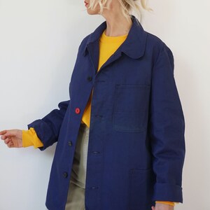 Pu&ograve; includere: Giacca da lavoro blu navy con chiusura a bottoni e una singola tasca sul petto. La giacca ha un colletto classico e maniche lunghe. Il polsino &egrave; arrotolato, rivelando una camicia gialla sotto. Un bottone &egrave; rosso.