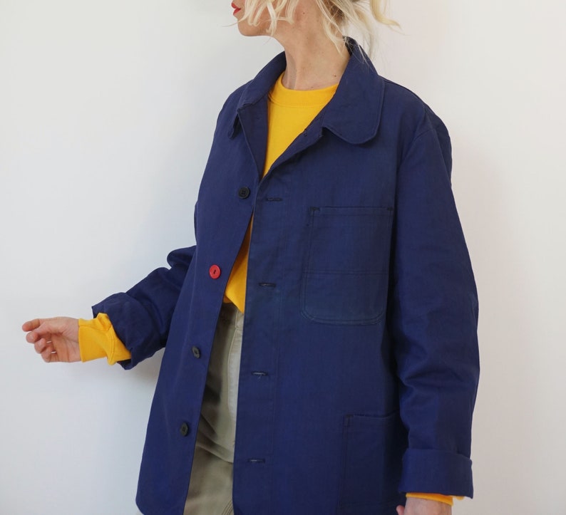 Pu&ograve; includere: Giacca da lavoro blu navy con chiusura a bottoni e una singola tasca sul petto. La giacca ha un colletto classico e maniche lunghe. Il polsino &egrave; arrotolato, rivelando una camicia gialla sotto. Un bottone &egrave; rosso.