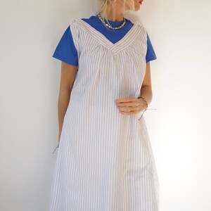 May include: A white and blue striped sleeveless dress with a V-neckline. The dress is worn over a blue t-shirt.