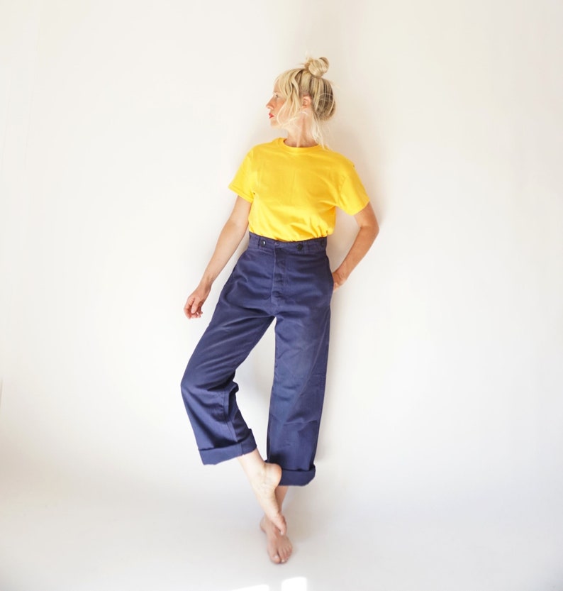 K&ouml;nnte beinhalten: Eine Frau tr&auml;gt ein gelbes T-Shirt und eine blaue, hochgeschlossene Hose mit umgeschlagenen Beinen. Sie steht auf einem Bein, das andere Bein ist im Knie gebeugt.