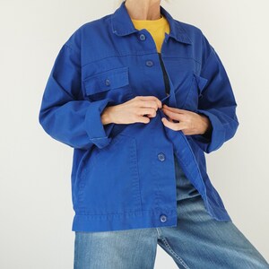 May include: A vibrant blue work jacket with a button-up front, two chest pockets, and two lower pockets. The jacket is paired with blue jeans and a yellow shirt underneath. The jacket has a classic collar and long sleeves.