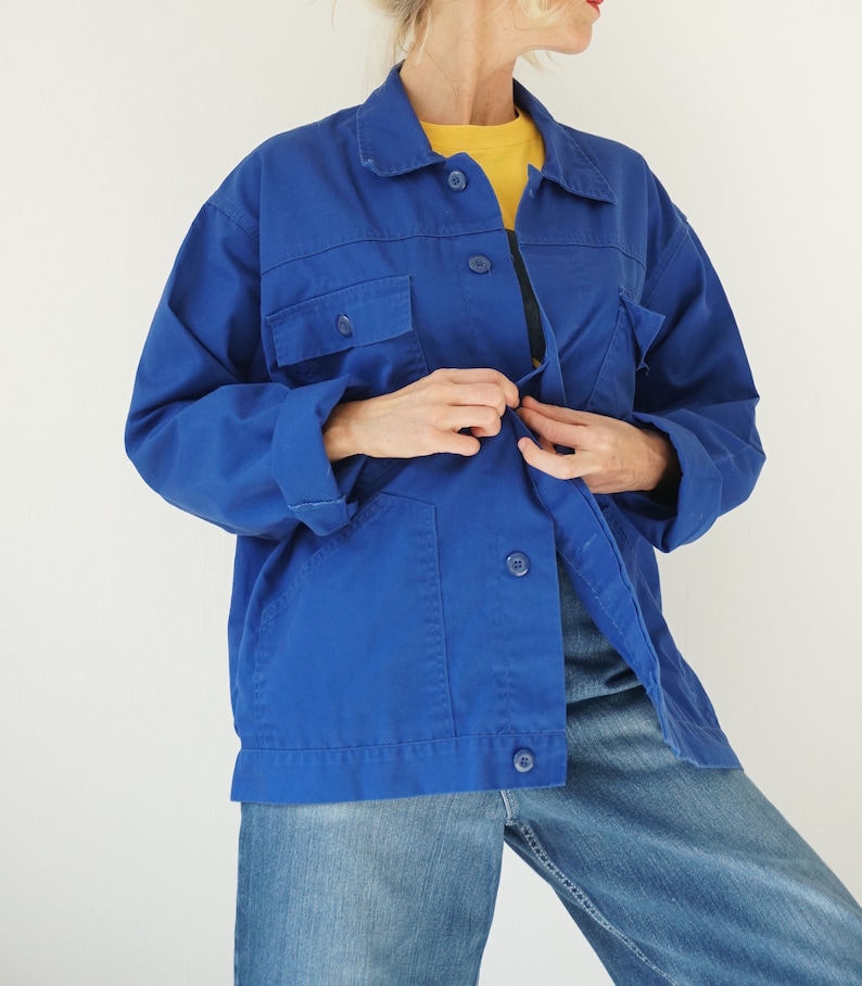 May include: A vibrant blue work jacket with a button-up front, two chest pockets, and two lower pockets. The jacket is paired with blue jeans and a yellow shirt underneath. The jacket has a classic collar and long sleeves.