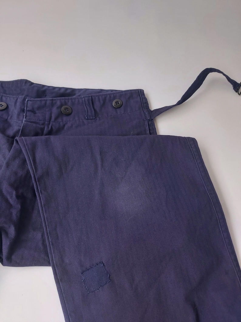 K&ouml;nnte beinhalten: Eine dunkelblaue Hose mit einem Flicken am rechten Bein. Die Hose hat einen Knopfverschluss und G&uuml;rtelschlaufen.
