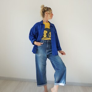 May include: A woman wearing a blue jacket over a yellow t-shirt and blue jeans. The t-shirt has a graphic of a dog and cat. The jeans are cuffed at the ankles.