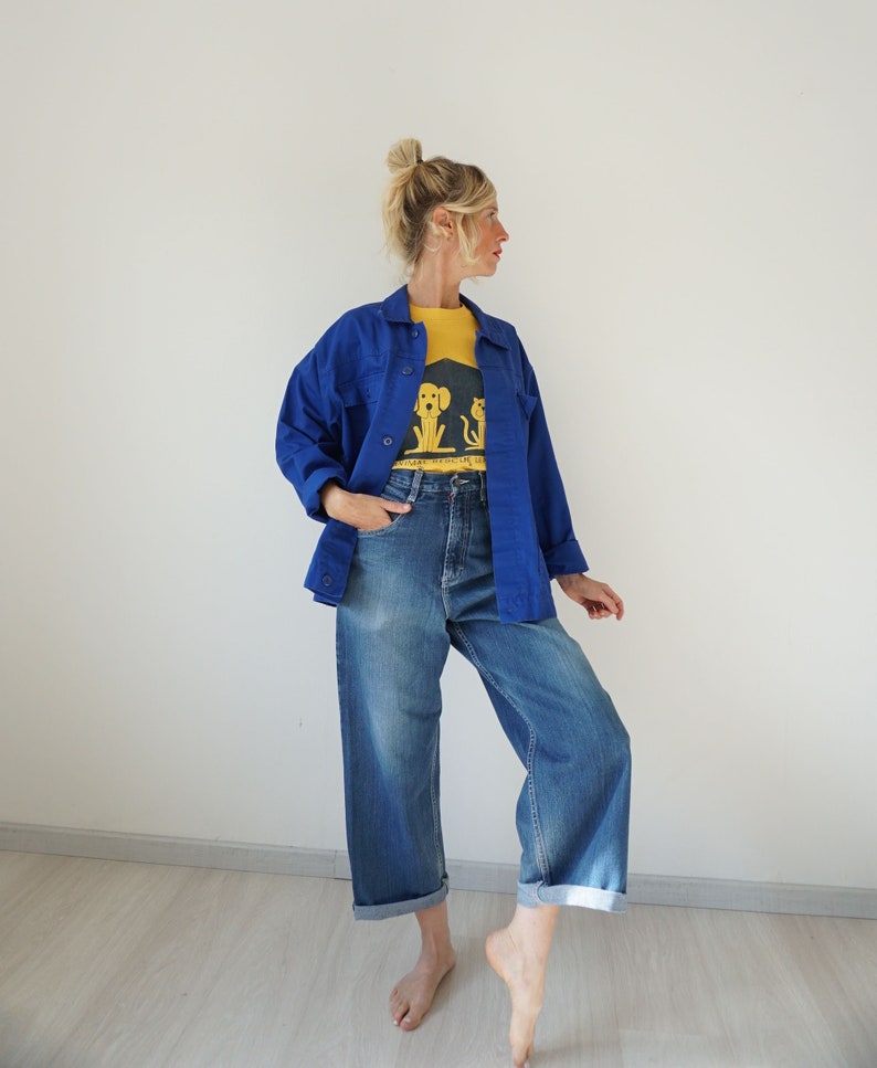 May include: A woman wearing a blue jacket over a yellow t-shirt and blue jeans. The t-shirt has a graphic of a dog and cat. The jeans are cuffed at the ankles.