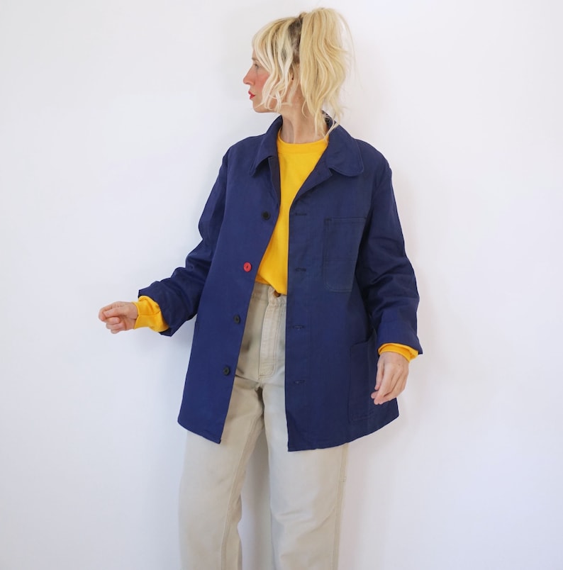 Pu&ograve; includere: Giacca blu navy con bottoni, un bottone rosso e due tasche anteriori. La giacca &egrave; indossata sopra una maglietta gialla a maniche lunghe e abbinata a pantaloni beige. La modella indossa la giacca.
