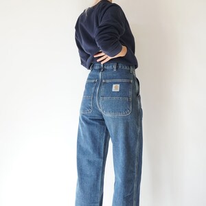 May include: Blue denim work pants with a straight leg cut. The pants have large back pockets and a small logo patch. The person in the image is wearing a navy blue sweatshirt.