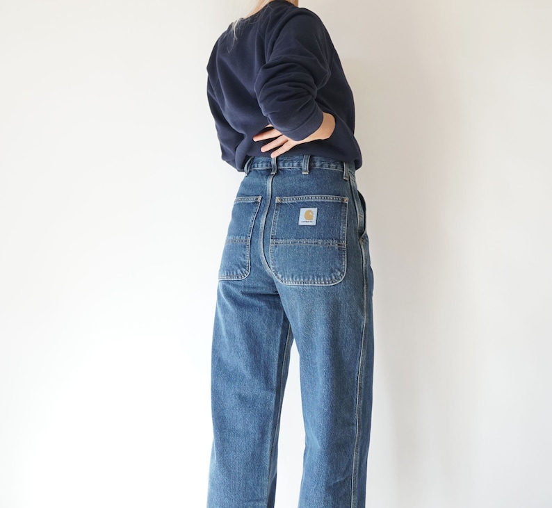 May include: Blue denim work pants with a straight leg cut. The pants have large back pockets and a small logo patch. The person in the image is wearing a navy blue sweatshirt.