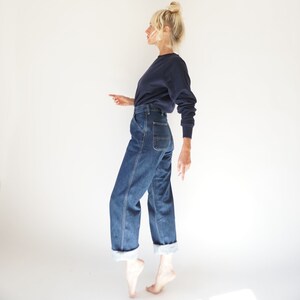 May include: A person wearing a navy blue long-sleeve shirt and blue denim jeans. The jeans have a straight leg with a folded cuff. The person is standing on their toes against a white background.