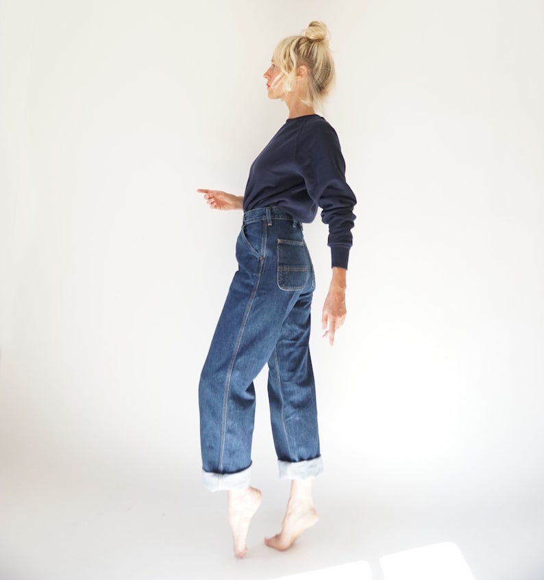 May include: A person wearing a navy blue long-sleeve shirt and blue denim jeans. The jeans have a straight leg with a folded cuff. The person is standing on their toes against a white background.