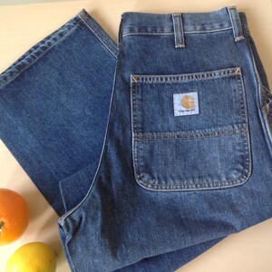 May include: Blue denim work pants with a Carhartt label on the back pocket. The pants are folded on a light-colored surface, with an orange and a lemon nearby. The pants have multiple pockets and a classic workwear design.