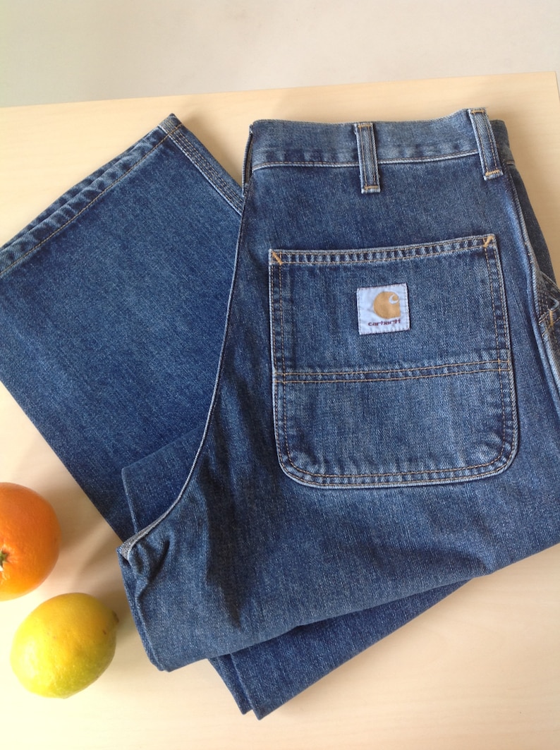 May include: Blue denim work pants with a Carhartt label on the back pocket. The pants are folded on a light-colored surface, with an orange and a lemon nearby. The pants have multiple pockets and a classic workwear design.