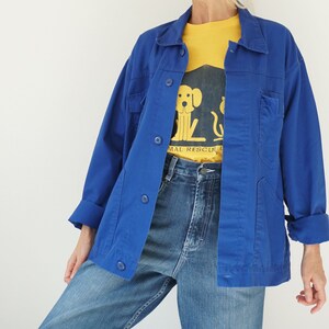 May include: A bright blue jacket with button closures and pockets, worn over a yellow t-shirt with a dog graphic and the words "ANIMAL RESCUE". The outfit includes blue jeans.
