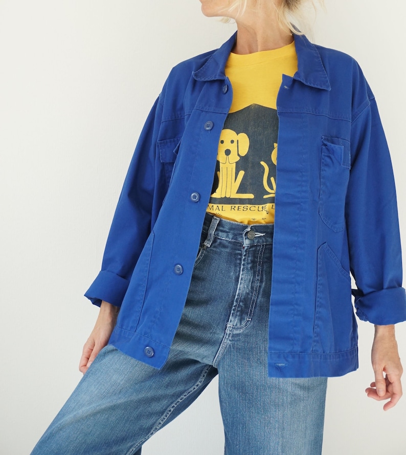 May include: A bright blue jacket with button closures and pockets, worn over a yellow t-shirt with a dog graphic and the words "ANIMAL RESCUE". The outfit includes blue jeans.