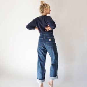 May include: A navy blue long-sleeve sweatshirt and blue denim jeans. The jeans have a straight leg, cuffed hem, and a Carhartt logo patch on the back pocket. The model is standing in front of a white background.