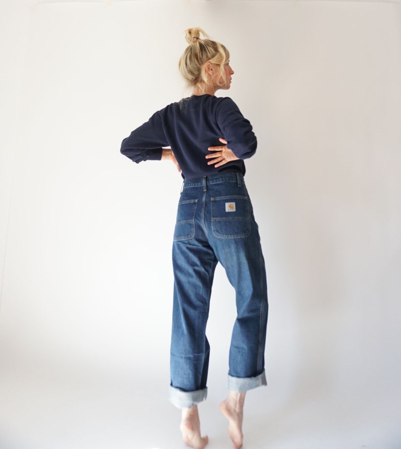 May include: A navy blue long-sleeve sweatshirt and blue denim jeans. The jeans have a straight leg, cuffed hem, and a Carhartt logo patch on the back pocket. The model is standing in front of a white background.