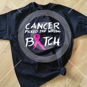 Puede incluir: Camiseta negra con texto blanco que dice "Cancer picked the wrong b*tch" con un gráfico de cinta rosa.