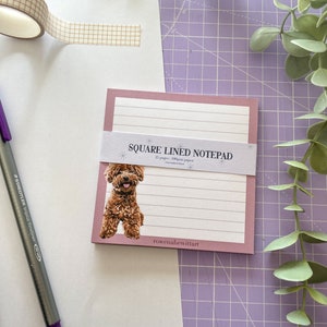 May include: A pink square lined notepad with a brown dog illustration on the front. The notepad has the text "SQUARE LINED NOTEPAD" and "rowenaewittart" printed on the front.