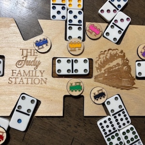Puede incluir: Un juego de dominó de madera con una base de madera cortada a medida que dice "THE Judy FAMILY STATION". El juego incluye fichas de dominó y fichas con temática de trenes. Los dominós son blancos con puntos negros.