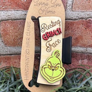 May include: Hand-painted hair claw with a wooden design. The claw features the text "Resting Grinch Face" in red and black, and a green Grinch face. It is attached to a brown card with the text "Lorna Lou Home".