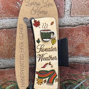 Könnte beinhalten: Handbemalte Haarspange mit Holzdesign. Die Spange zeigt eine Kaffeetasse, Herbstblätter und einen Schal mit den Worten "Sweater Weather". Die Spange ist an einer braunen Kartonrückseite mit dem Text "Lorna Lou Home" befestigt.