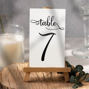 May include: A white card with the word "table" in elegant script and the number "7" in bold black font. The card is displayed on a small wooden easel, perfect for weddings or events.