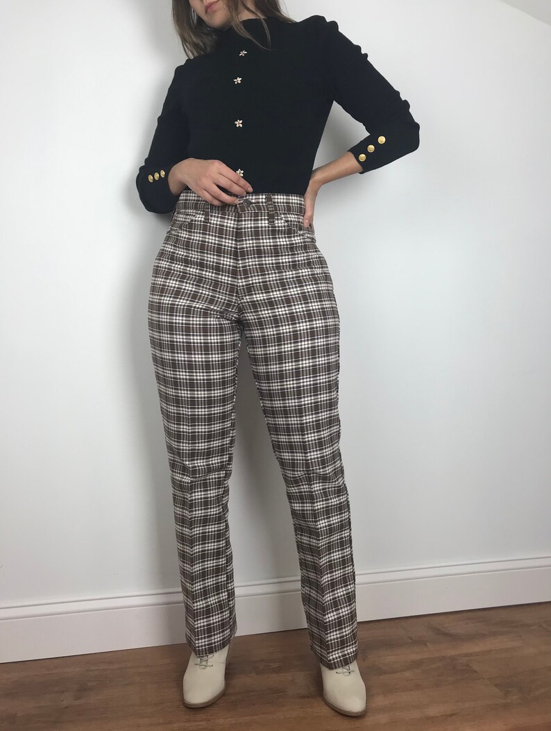 May include: A black mock neck sweater with gold buttons on the front and cuffs. The sweater is paired with brown and white plaid pants.