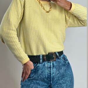 May include: A yellow sweater with a subtle blue pattern is paired with acid wash jeans and a dark green belt with a decorative buckle. A gold chain necklace adds a touch of elegance to the outfit.