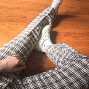 May include: A person wearing brown and white plaid pants with white Converse sneakers. The sneakers have a black star on the side.