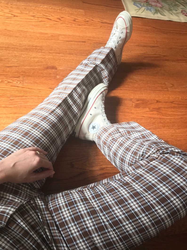 May include: A person wearing brown and white plaid pants with white Converse sneakers. The sneakers have a black star on the side.