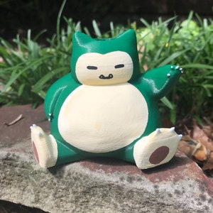 Peut inclure: Une figurine de Snorlax verte et jaune avec un ventre blanc et des pieds bruns. La figurine est assise sur un rocher.