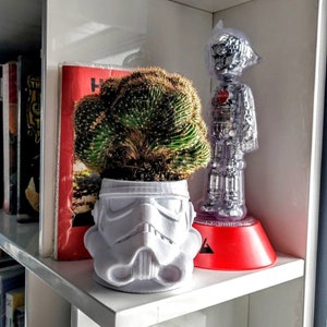 Puede incluir: Una maceta de plástico blanco con forma de casco de Stormtrooper de Star Wars, que contiene una planta de cactus verde. La maceta está en un estante blanco con una base roja y una figura de robot de plástico transparente.
