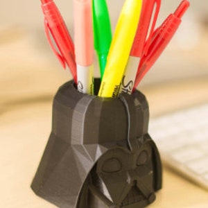May include: A black 3D printed Darth Vader head pen holder with five pens inserted. The pens are red, pink, green, yellow, and red.