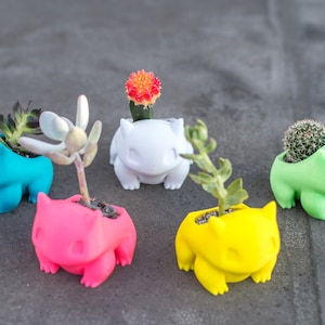 May include: Five small ceramic planters shaped like a cartoon character, each with a different coloured glaze. The planters are filled with small succulents and cacti.