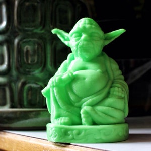 May include: Green Yoda figurine sitting in a meditative pose, with a green base. The figurine is made of a smooth, plastic-like material.