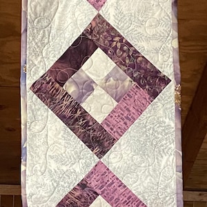May include: A white table runner with a quilted design featuring three large purple diamond shapes. The diamonds are made of different purple fabrics with floral and geometric patterns.