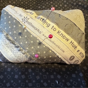May include: A gray and white fabric pincushion with a label that reads "Getting to Know Hue #9718 for Clothworks www.clothworks.com". The pincushion has several pins with colored heads sticking out of it.