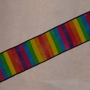 Puede incluir: Un camino de mesa largo y estrecho con rayas de arcoíris y bordes negros. El camino de mesa presenta rayas alternas de rojo, naranja, amarillo, verde, azul, morado y rosa.