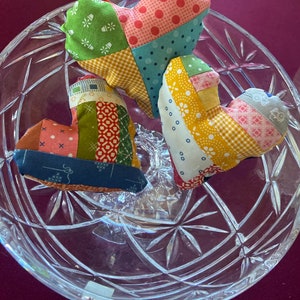 May include: Three patchwork fabric heart-shaped cushions in shades of red, green, yellow, blue, and pink. The cushions are displayed on a clear glass pedestal.