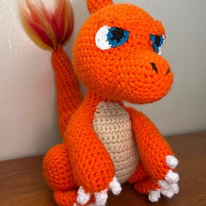 May include: A handmade orange crochet Charmander plush toy with white accents on its feet and eyes. The tail has a flame-like design with yellow and red yarn. The belly is a light tan color.